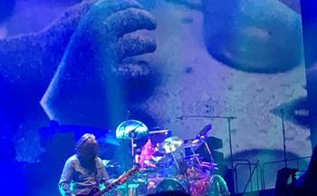 Tool guitarist playing in front of the drummer, illuminated in a blue glow.