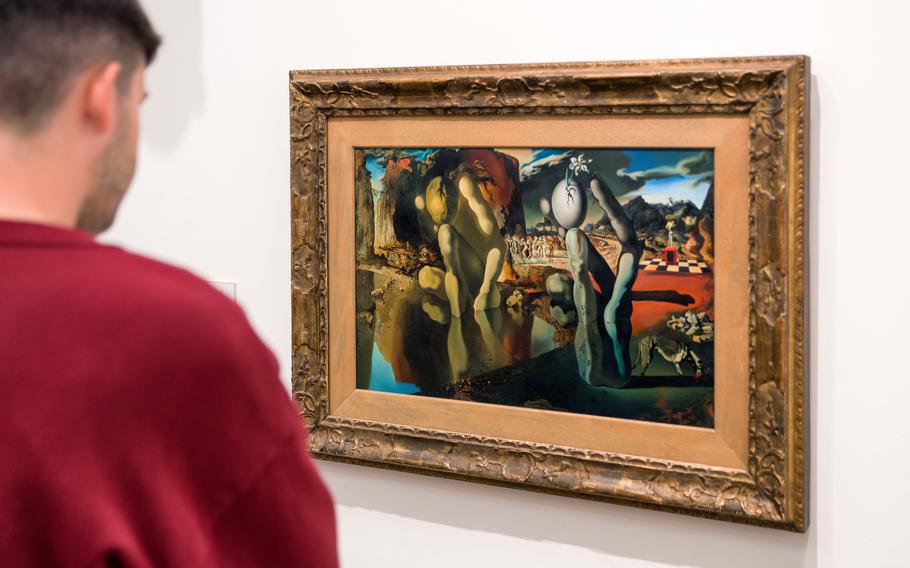 A man in a red shirt looking at a colorful representation painting of two mannequin-like figures dipicting Narcissus. 