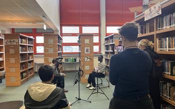 a student films a video interview and two other faculty members sit in a library looking on. 