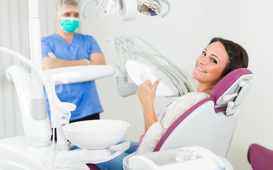 Woman facing camera, sitting back in a dental chair. She is smiling and giving a “thumbs up” symbol. Behind her, a dentist has his arms folded and looking at patient.