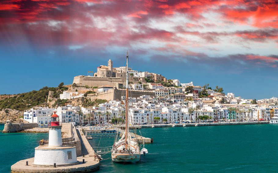 Scenic view of Ibiza Port, showcasing luxury yachts docked along the waterfront, historic buildings of Dalt Vila rising in the background, and a lively promenade with shops and cafes.