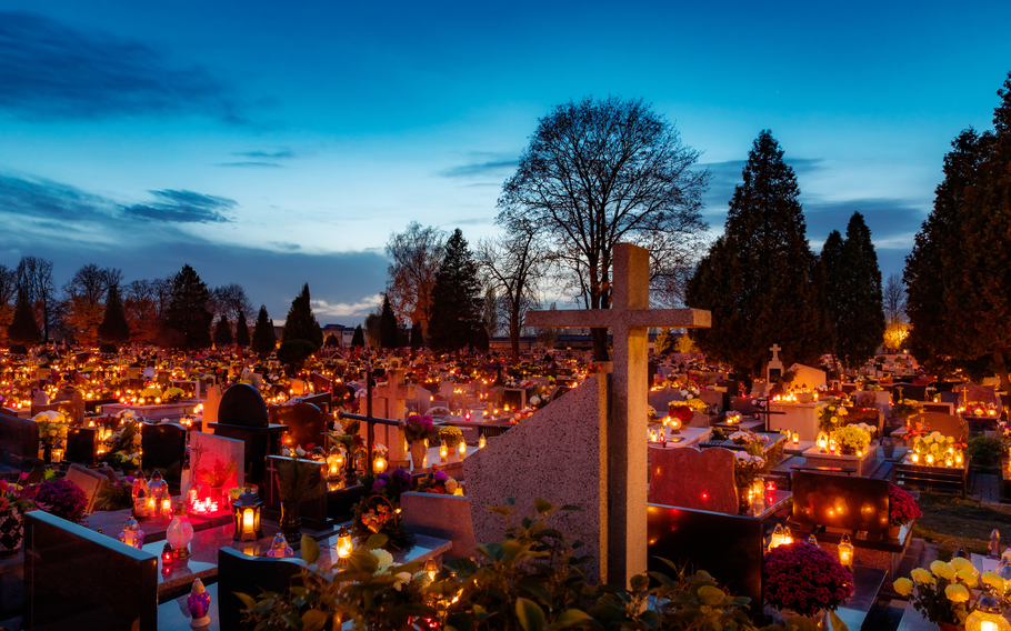 A cemetery at dusk with rows of graves adorned with lit candles and fresh flowers, creating a tranquil and reflective scene. The soft glow of the candles contrasts with the darkening blue sky, and tall trees outline the background, adding to the solemn and peaceful atmosphere of All Saints’ Day. A stone cross in the foreground stands as a prominent symbol of remembrance.