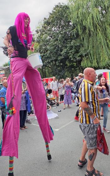 A person in bright pink pants with bright pink hair standing tall on stilts with other people surrounding them for the Pride parade