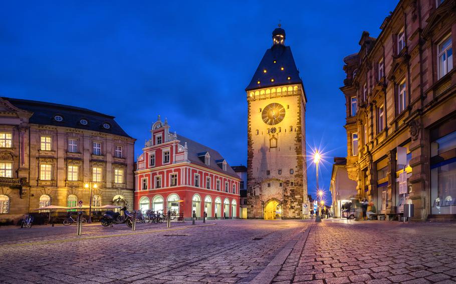 This enchanting nighttime view captures the historic charm of a European city square, illuminated by warm streetlights against a deep blue sky. The centerpiece is a striking clock tower with glowing details, flanked by beautifully preserved buildings, including a vibrant red structure with intricate architecture. The cobblestone streets add to the old-world ambiance, inviting visitors to explore and soak in the atmosphere.