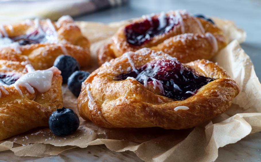 This image showcases freshly baked blueberry danishes with golden, flaky pastry layers filled with glossy blueberry compote. The pastries are drizzled with a light glaze, dusted with powdered sugar, and surrounded by fresh blueberries for a perfect balance of sweetness and texture.