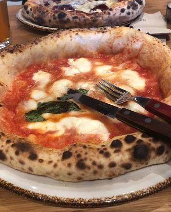 Napoli-style pizza with thick, wood-fired crust. Pizza features tomato-based sauce, with blobs of mozzarella scattered throughout, and basil leaves at the center. There is a knife and fork on top of the pizza.