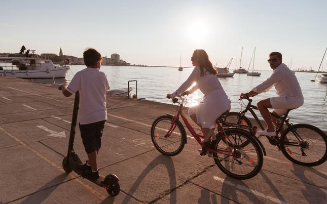 Pre-teen boy facing away from camera on electric scooter riding next to his mom and dad who are both riding bikes. They are all riding next to blue water which has several sailboats on it.