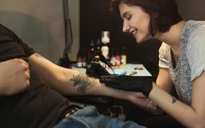 Cheerful woman tattooist doing tattoo with tattoo machine on male arm. 