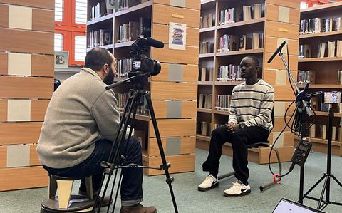 Photo Of A man sits in a library looking into a video camera as he sets up for an interview with a student. 