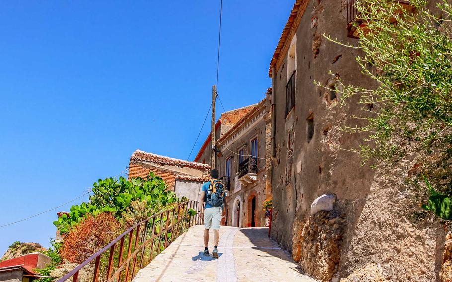 A hiker walks through the narrow, sunlit streets of the ancient village of Pentedattilo, Italy, surrounded by rustic stone houses and scenic Mediterranean views.