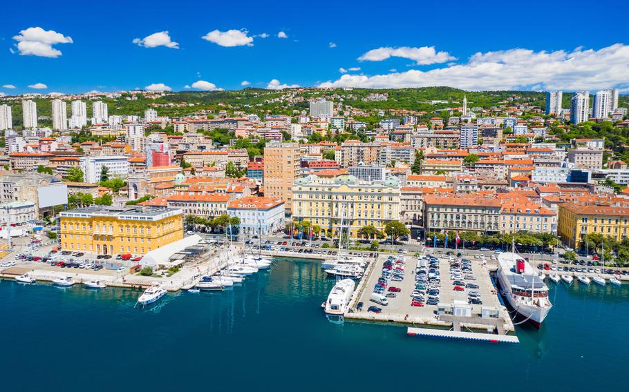 A vibrant aerial view of Rijeka, Croatia, showcasing its colorful waterfront buildings, bustling marina with docked yachts, and lush green hills in the background under a bright blue sky with scattered white clouds.