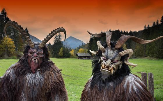 Photo Of Two people in Krampus costumes with a Alpine/mountain background. The Krampuses are covered in brown, white, and black fur. The Krampus on the left has two long, curled horns and the Krampus on the right has four long, curled horns. The Krampus on the left has one long fang among a row of sharp teeth. The Krampus on the left has four long fangs, two facing up, two facing down among rows of sharp teeth.