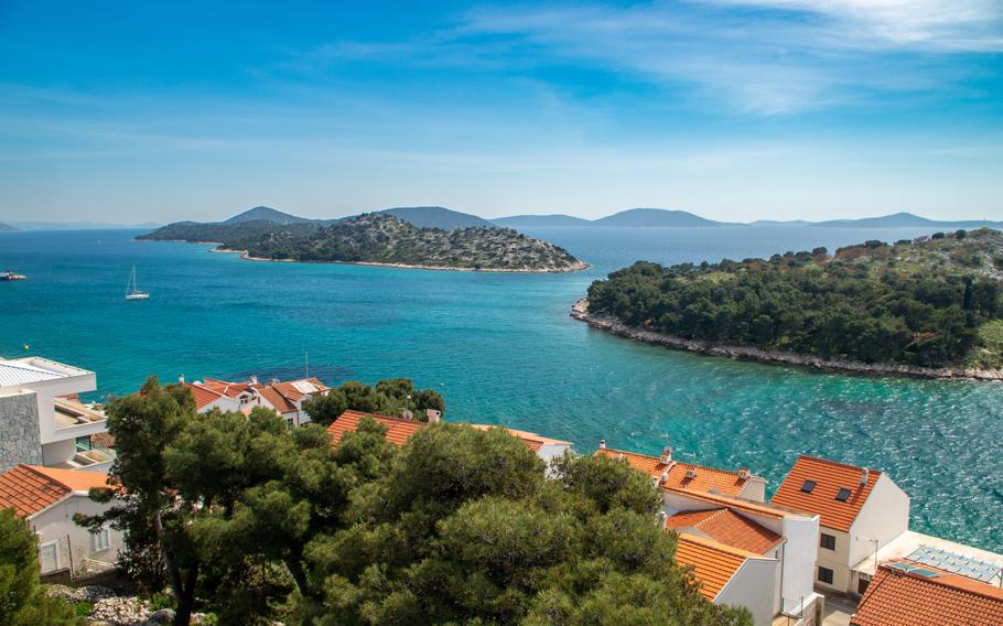 An idyllic coastal view of Croatia, featuring turquoise waters, small islands covered with greenery, and orange-roofed houses nestled among trees in the foreground, all under a clear blue sky.