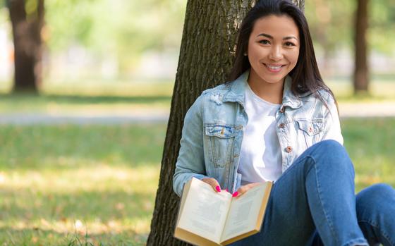 Woman with a book in the park. She sits on the grass with a book, looking at camera and smiling. 