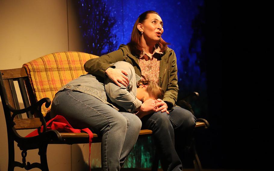 Two people sitting on a bench on stage. One woman is singing looking up into the sky; the other has their head in the lap of the person singing as if in anguish.