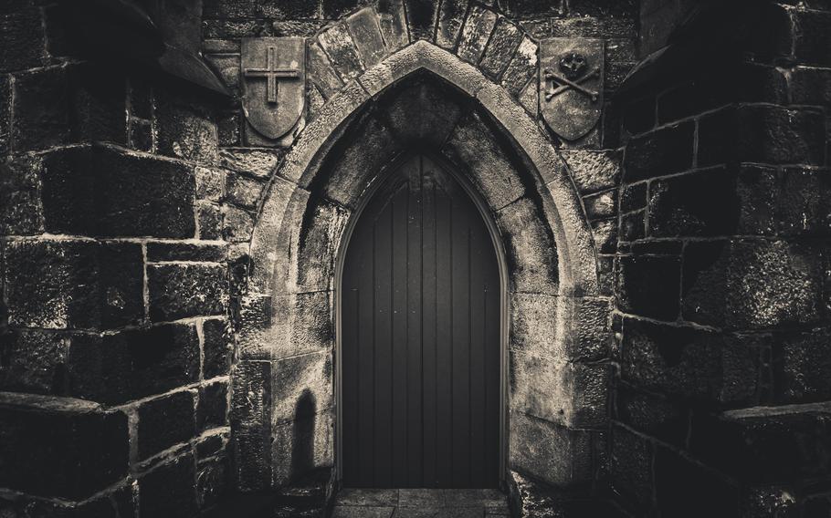 A dark, gothic castle entrance framed by heavy stone walls, featuring eerie carvings of a cross and skull, evoking a sense of mystery and foreboding.