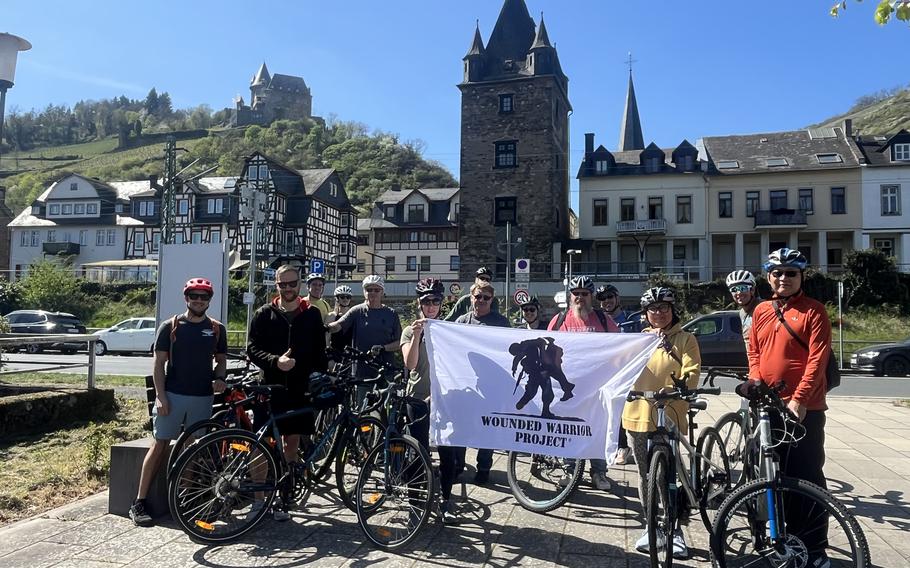 A group of cyclists stand beside their bikes holding a Wounded Warrior Project banner in front of a medieval German town.