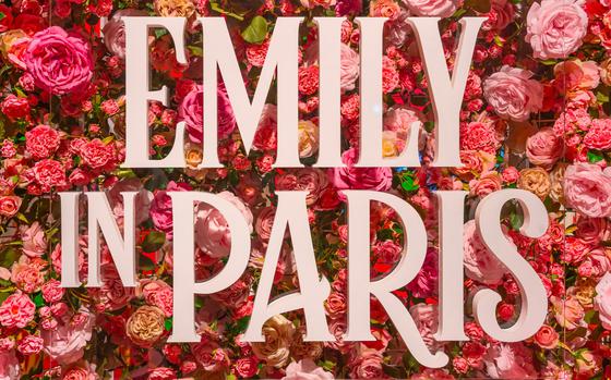The title “Emily in Paris” in bright pink letters hanging in front of a backdrop of bright red and pink flowers like roses and peonies. 