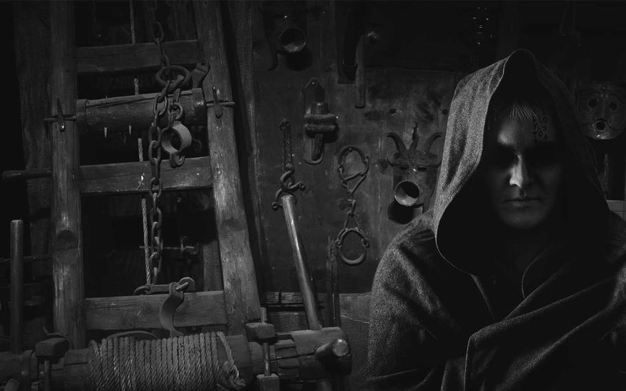 A hooded figure sits ominously in a dark room surrounded by an array of medieval torture tools, creating a chilling and foreboding atmosphere.