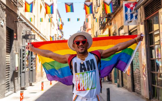 Man standing in the middle of an empty alleyway under a string of pride flags, holding a large Pride flag behind him with both hands. He is wearing a white tank top which reads, “I AM PROUD,” with the word “proud” featuring a rainbow background. He is also wearing white pants with rainbow suspenders next to the pockets. He is wearing red sunglasses with the rims shaped like hearts. On top of his head is a white hat.