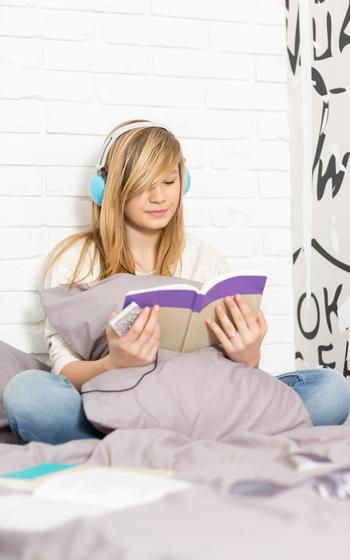 Teenage girl listening to music while reading book in bedroom
