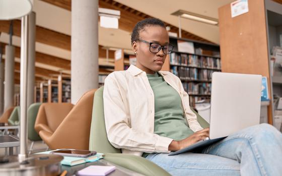 Busy short-haired African girl student using laptop technology sitting in university campus library. Serious young Black woman elearning looking at computer advertising hybrid work and elearning.
