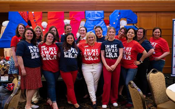 Group photo of 15 people wearing a variety of red, white and blue shirts reading “I’m in my AWAG era” against red and blue sign reading AWAG