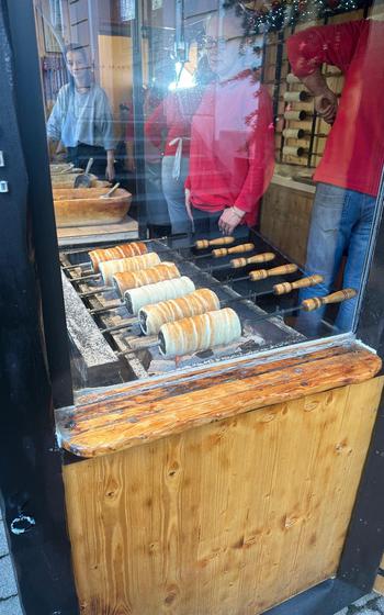 Cylindrical dough on skewers turning golden brown over hot, red coals. 