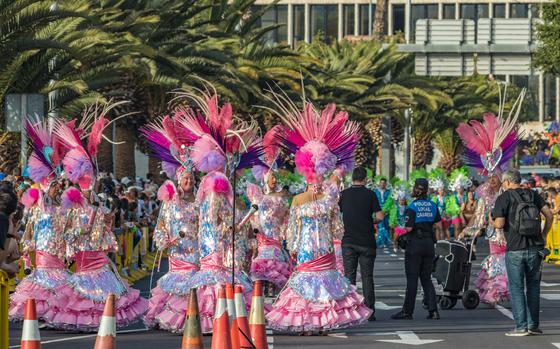 a colorful picture of people dressed in pink gittering costumes for Carnival, palm trees are in the background and there are people lining the streets.