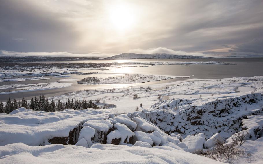 A white snow-covered landscape that includes a lake and mountains in the background with a few pine trees poking up out of the snow.