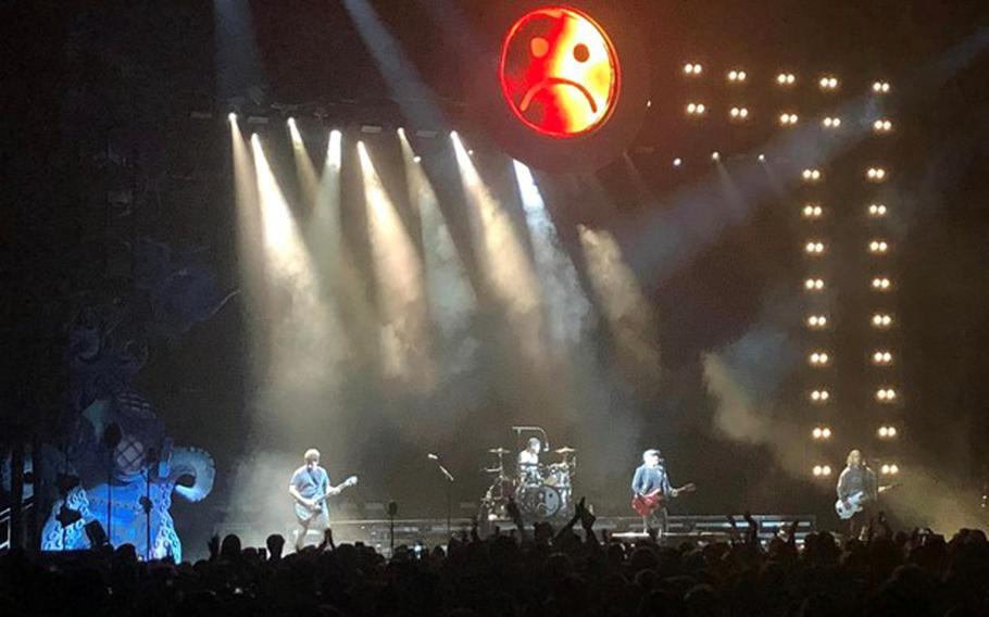 Seeing crowd with arms in the air, then,  on stage (from left to right) guitarist, drummer, lead singer and bassist.  From above, stage lights are shining down onto the band. From above, center stage: the album symbol, half sad-face and half smiling-face, lit above the stage.