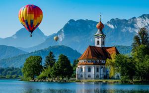 A beautiful red and blue hot-air ballon floats above bright blue water. There are mountains in the background and a beautiful little church. 