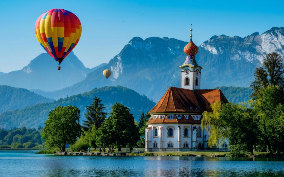 A beautiful red and blue hot-air ballon floats above bright blue water. There are mountains in the background and a beautiful little church. 