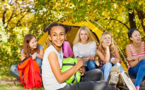 Happy African girl holding green sleeping bag sitting near at camping place with her friends