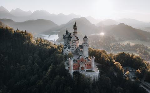 Photo Of A view of a fairytale like castle thats white and brown on top of a hill with trees and mountains in the background 