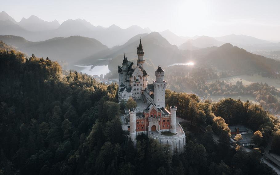 A view of a fairytale like castle thats white and brown on top of a hill with trees and mountains in the background 