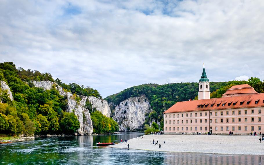A red tile roofed monastery sits along a beach at the edge of the Danube River, with cliffs lining the opposite shore. 