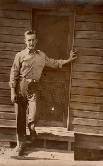 an old sepia-toned photograph of a young soldier at fort Stewart leaning against an old wooden house