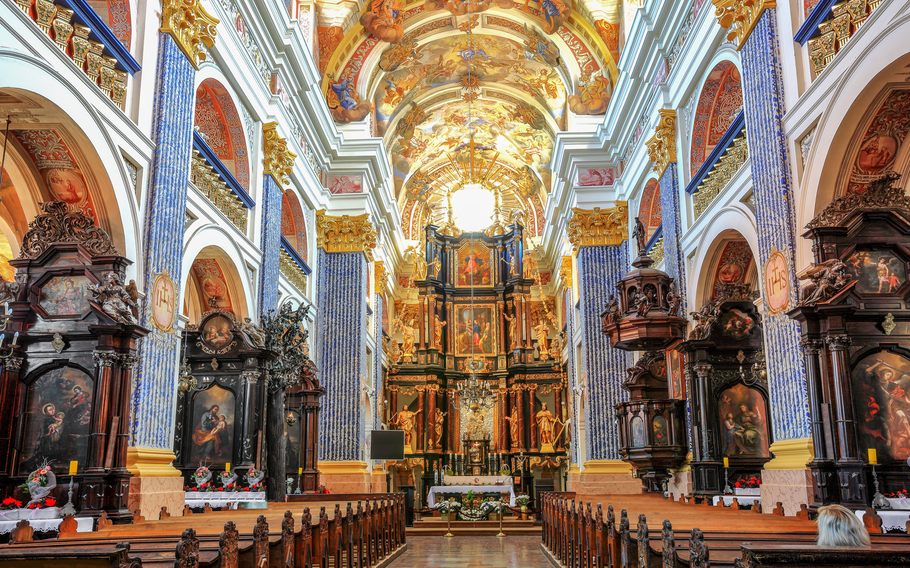 The interior of a grand church featuring tall, elaborately decorated columns, vibrant frescoes depicting religious scenes on the arched ceiling, and intricate wood carvings. The main altar is richly adorned with statues, golden accents, and detailed paintings, creating a majestic focal point. Rows of wooden pews lead towards the altar, inviting reflection and prayer in the serene and awe-inspiring space.