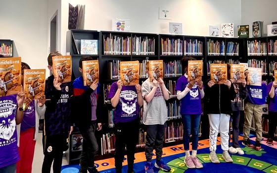 Eleven kids line up in front of library shelves and hold a bright yellow Dandelions magazine in front of their faces 