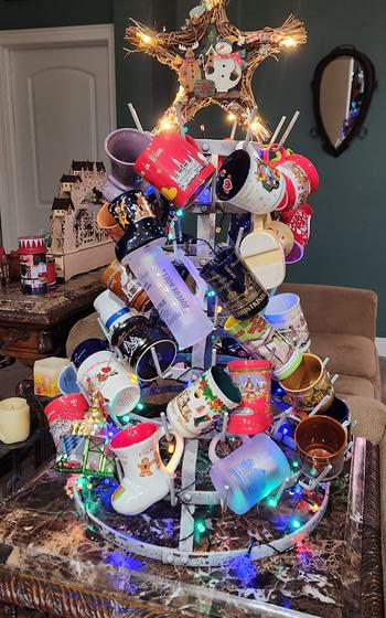 A metal Christmas tree with hooks displays many different shaped Christmas market mugs on a wooden table. There’s a gold star atop the tree and lots of red and white mugs. 