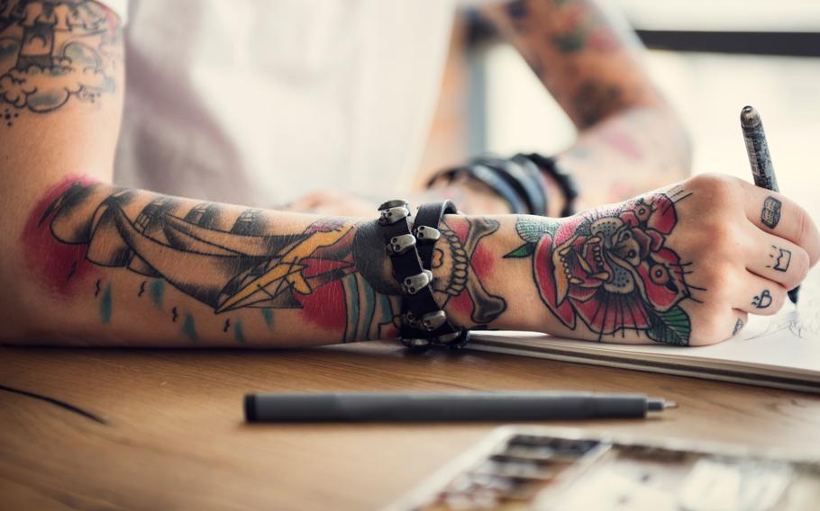 Arm with sailboat tattoo and hand with rose tattoo holding pencil