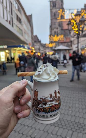 a hand holds out a white Christmas mug with a snowy village on it. It is filled with whipped-cream and a cinnamon stick. 