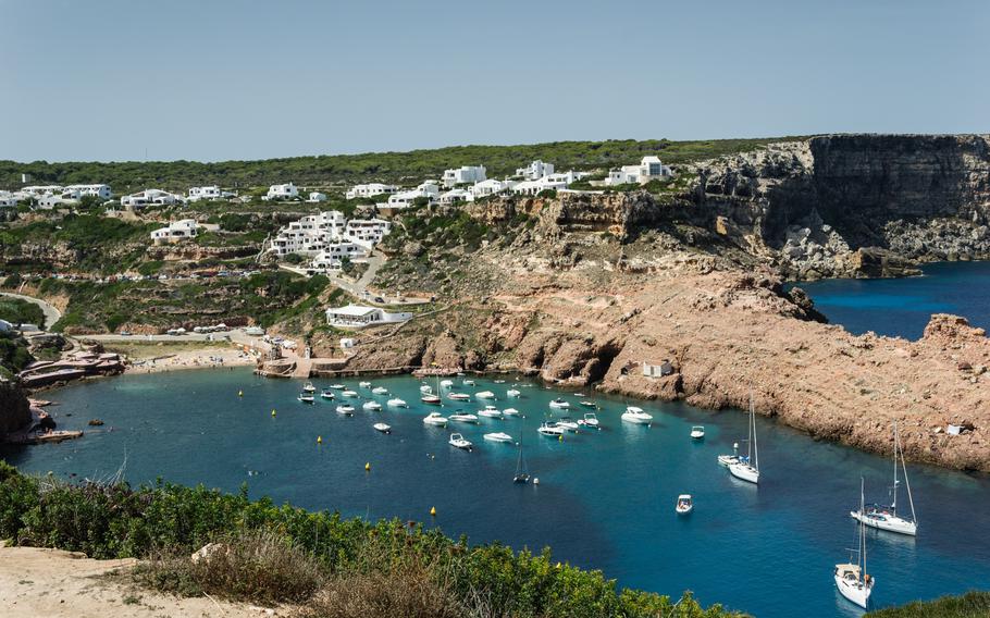 Idyllic view of Cala Morell in Menorca, featuring crystal-clear turquoise waters surrounded by rugged cliffs and lush greenery, with prehistoric caves nestled into the rocky landscape.