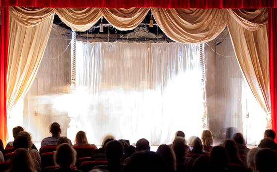 Defocused silhouettes of people on stage in the theater. Point of view of reader behind audience members looking at the stage.