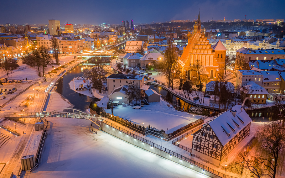 Bydgoszcz, Poland, captured in winter at night, showcasing a snowy cityscape with a blend of historic and modern architecture. The illuminated Gothic church and half-timbered buildings stand out, with softly lit bridges spanning the Brda River, creating a serene and enchanting atmosphere.