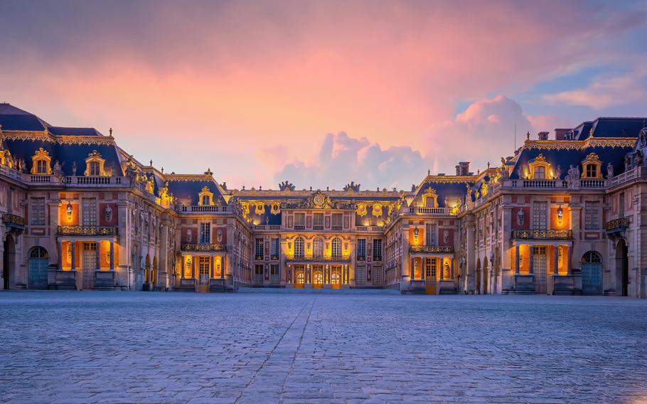 The enchanting entrance of the Château de Versailles, near Paris, France, captured at sunset. The palace’s iconic architecture is bathed in warm golden light, accentuating its intricate details, gilded accents, and majestic symmetry. The soft hues of the sunset, blending pink and orange, create a dreamy backdrop for this historic masterpiece. The vast cobblestone courtyard in the foreground leads visitors toward the grandeur of one of France’s most celebrated landmarks.