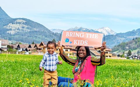 Photo Of Woman with toddler with mountains in the background | Woman is holding a sign that reads “Bring the Kids”