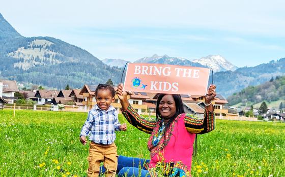 Woman with toddler with mountains in the background | Woman is holding a sign that reads “Bring the Kids”