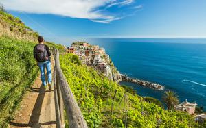 A person with a backpack hikes through a grassy trail high on a cliffside. the colorful Cinque Terre houses are up ahead in the distance above a bright blue ocean. 